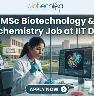 MSc Biotechnology & Biochemistry Job at IIT Delhi – Enzyme Assay Specialist Role Open Now! MSc Biotechnology & Biochemistry Job at IIT Delhi. A professional and realistic featured image for a job opening. At the top center is the Biotecnika Global logo, followed by large, bold text reading "MSc Biotechnology & Biochemistry Job at IIT Delhi." The central scene depicts a female researcher in a white lab coat working in a high-tech laboratory, using a pipette on colorful samples while viewing a 3D molecular structure on a large monitor. The background shows advanced lab equipment and a clear view of the IIT Delhi Research campus through large windows. At the bottom, there is a prominent teal button that says "APPLY NOW" with a white arrow icon.