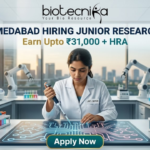 NIPER Ahmedabad Hiring Junior Research Fellow Earn Upto ₹31,000 + HRA Apply Fast! A professional featured image for a Junior Research Fellow (JRF) job opening at NIPER Ahmedabad, created for Biotecnika. The scene features a female scientist in a white lab coat using a pipette over a glowing, futuristic digital workbench displaying molecular structures. The laboratory is equipped with advanced technology, including robotic arms and high-end centrifuges, with a modern city skyline visible through large windows in the background. The bold heading "NIPER AHMEDABAD HIRING JUNIOR RESEARCH FELLOW" and the tagline "Earn Upto ₹31,000 + HRA" are prominently displayed, with an "Apply Now" button at the bottom.