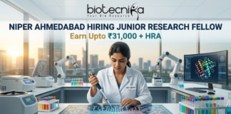 NIPER Ahmedabad Hiring Junior Research Fellow Earn Upto ₹31,000 + HRA Apply Fast! A professional featured image for a Junior Research Fellow (JRF) job opening at NIPER Ahmedabad, created for Biotecnika. The scene features a female scientist in a white lab coat using a pipette over a glowing, futuristic digital workbench displaying molecular structures. The laboratory is equipped with advanced technology, including robotic arms and high-end centrifuges, with a modern city skyline visible through large windows in the background. The bold heading "NIPER AHMEDABAD HIRING JUNIOR RESEARCH FELLOW" and the tagline "Earn Upto ₹31,000 + HRA" are prominently displayed, with an "Apply Now" button at the bottom.
