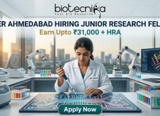 NIPER Ahmedabad Hiring Junior Research Fellow Earn Upto ₹31,000 + HRA Apply Fast! A professional featured image for a Junior Research Fellow (JRF) job opening at NIPER Ahmedabad, created for Biotecnika. The scene features a female scientist in a white lab coat using a pipette over a glowing, futuristic digital workbench displaying molecular structures. The laboratory is equipped with advanced technology, including robotic arms and high-end centrifuges, with a modern city skyline visible through large windows in the background. The bold heading "NIPER AHMEDABAD HIRING JUNIOR RESEARCH FELLOW" and the tagline "Earn Upto ₹31,000 + HRA" are prominently displayed, with an "Apply Now" button at the bottom.