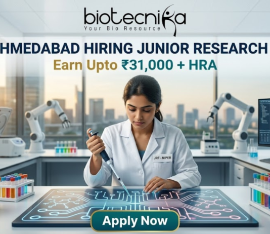 NIPER Ahmedabad Hiring Junior Research Fellow | Earn Upto ₹31,000 + HRA | Apply Fast! NIPER Ahmedabad Hiring Junior Research Fellow Earn Upto ₹31,000 + HRA Apply Fast! A professional featured image for a Junior Research Fellow (JRF) job opening at NIPER Ahmedabad, created for Biotecnika. The scene features a female scientist in a white lab coat using a pipette over a glowing, futuristic digital workbench displaying molecular structures. The laboratory is equipped with advanced technology, including robotic arms and high-end centrifuges, with a modern city skyline visible through large windows in the background. The bold heading "NIPER AHMEDABAD HIRING JUNIOR RESEARCH FELLOW" and the tagline "Earn Upto ₹31,000 + HRA" are prominently displayed, with an "Apply Now" button at the bottom.