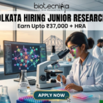 NIPER Kolkata Hiring Junior Research Fellow. Alt Text: A high-quality, professional featured image for a "Junior Research Fellow" job opening at NIPER Kolkata. The scene features a focused female scientist in a white lab coat and safety glasses working in a futuristic laboratory. Through the large windows, iconic Kolkata landmarks like the Victoria Memorial and the Howrah Bridge are visible at sunset. The image includes the Biotecnika logo at the top center, the bold headline "NIPER KOLKATA HIRING JUNIOR RESEARCH FELLOW," and a tagline stating "Earn Upto ₹37,000 + HRA." A prominent "APPLY NOW" button is positioned at the bottom center.