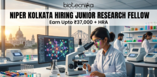 NIPER Kolkata Hiring Junior Research Fellow | Get Upto ₹37,000 + HRA | Apply Online Now!! NIPER Kolkata Hiring Junior Research Fellow. Alt Text: A high-quality, professional featured image for a "Junior Research Fellow" job opening at NIPER Kolkata. The scene features a focused female scientist in a white lab coat and safety glasses working in a futuristic laboratory. Through the large windows, iconic Kolkata landmarks like the Victoria Memorial and the Howrah Bridge are visible at sunset. The image includes the Biotecnika logo at the top center, the bold headline "NIPER KOLKATA HIRING JUNIOR RESEARCH FELLOW," and a tagline stating "Earn Upto ₹37,000 + HRA." A prominent "APPLY NOW" button is positioned at the bottom center.
