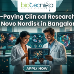 Novo Nordisk Hiring Clinical Data Scientist – High-Paying Clinical Research Job in Bangalore! A promotional featured image for a job opening, featuring the headline "High-Paying Clinical Research Job at Novo Nordisk in Bangalore!" in bold white text at the top. The Biotecnika logo is positioned at the top center. The central visual shows two professional researchers—a woman and a man in white lab coats—smiling and collaborating while looking at a digital tablet that displays a glowing neural network. They are standing in a futuristic, high-tech laboratory equipped with large, curved monitors showing 3D protein structures and complex biological data. Through the large glass windows behind them, a modern city skyline is visible during a warm sunset. At the bottom center, there is a prominent, stylized "APPLY NOW" button.
