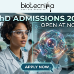 PhD Admissions 2026 Open at NCCS Pune Life Science Graduates Must Apply Before Deadline! A professional and futuristic featured image for PhD Admissions 2026 at NCCS Pune. The image depicts a female researcher in a white lab coat and safety goggles interacting with a vibrant, glowing 3D holographic cell model that displays complex biological pathways. The setting is a modern, high-tech laboratory with other scientists working in the background. The Biotecnika logo is placed at the top center, the main heading reads "PhD ADMISSIONS 2026 OPEN AT NCCS PUNE," and a sleek "APPLY NOW" button with a cursor icon is located at the bottom center.