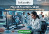 Project Assistant Job at National Institute of Immunology – Apply for Life Science Research Role Now! A promotional graphic for "biotecnika Your Bio Resource," set in a modern, busy laboratory environment. At the top center, the text "biotecnika Your Bio Resource" is displayed. Below it, the large headline "Project Assistant Job at National Institute of Immunology" is prominently featured in a white, sans-serif font. The main image shows a woman in a white lab coat, glasses, and blue gloves, using a pipette at a desk with dual computer monitors displaying complex molecular structures and scientific data. Other researchers work in the background. A large green button at the bottom contains the text "APPLY NOW". Various lab equipment is visible, and the overall color scheme is in blues, greens, white, and gold.