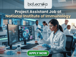 Project Assistant Job at National Institute of Immunology – Apply for Life Science Research Role Now! A promotional graphic for "biotecnika Your Bio Resource," set in a modern, busy laboratory environment. At the top center, the text "biotecnika Your Bio Resource" is displayed. Below it, the large headline "Project Assistant Job at National Institute of Immunology" is prominently featured in a white, sans-serif font. The main image shows a woman in a white lab coat, glasses, and blue gloves, using a pipette at a desk with dual computer monitors displaying complex molecular structures and scientific data. Other researchers work in the background. A large green button at the bottom contains the text "APPLY NOW". Various lab equipment is visible, and the overall color scheme is in blues, greens, white, and gold.