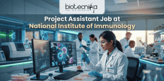 Project Assistant Job at National Institute of Immunology – Apply for Life Science Research Role Now! A promotional graphic for "biotecnika Your Bio Resource," set in a modern, busy laboratory environment. At the top center, the text "biotecnika Your Bio Resource" is displayed. Below it, the large headline "Project Assistant Job at National Institute of Immunology" is prominently featured in a white, sans-serif font. The main image shows a woman in a white lab coat, glasses, and blue gloves, using a pipette at a desk with dual computer monitors displaying complex molecular structures and scientific data. Other researchers work in the background. A large green button at the bottom contains the text "APPLY NOW". Various lab equipment is visible, and the overall color scheme is in blues, greens, white, and gold.