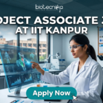 Project Associate Job at IIT Kanpur Life Science Research 2026. A promotional featured image banner for a 'PROJECT ASSOCIATE JOB AT IIT KANPUR', with the 'biotecnika YOUR BIO RESOURCE' logo at the top left. The image depicts a female scientist in a white lab coat and safety glasses, seated at a desk in a modern laboratory. She is interacting with a large, transparent holographic display, showing detailed 3D molecular structures and data visualizations. In the background, there is a large window with a view of the 'IIT KANPUR' campus building, other scientific equipment like microscopes and centrifuges, and a corkboard with certificates. A prominent, clickable 'Apply Now' button with a cursor arrow is located at the bottom center.