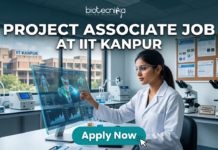 IIT Kanpur Hiring NOW! Project Associate Job Openings in Gene Therapy Project Associate Job at IIT Kanpur Life Science Research 2026. A promotional featured image banner for a 'PROJECT ASSOCIATE JOB AT IIT KANPUR', with the 'biotecnika YOUR BIO RESOURCE' logo at the top left. The image depicts a female scientist in a white lab coat and safety glasses, seated at a desk in a modern laboratory. She is interacting with a large, transparent holographic display, showing detailed 3D molecular structures and data visualizations. In the background, there is a large window with a view of the 'IIT KANPUR' campus building, other scientific equipment like microscopes and centrifuges, and a corkboard with certificates. A prominent, clickable 'Apply Now' button with a cursor arrow is located at the bottom center.