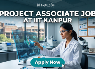 IIT Kanpur Hiring NOW! Project Associate Job Openings in Gene Therapy Project Associate Job at IIT Kanpur Life Science Research 2026. A promotional featured image banner for a 'PROJECT ASSOCIATE JOB AT IIT KANPUR', with the 'biotecnika YOUR BIO RESOURCE' logo at the top left. The image depicts a female scientist in a white lab coat and safety glasses, seated at a desk in a modern laboratory. She is interacting with a large, transparent holographic display, showing detailed 3D molecular structures and data visualizations. In the background, there is a large window with a view of the 'IIT KANPUR' campus building, other scientific equipment like microscopes and centrifuges, and a corkboard with certificates. A prominent, clickable 'Apply Now' button with a cursor arrow is located at the bottom center.
