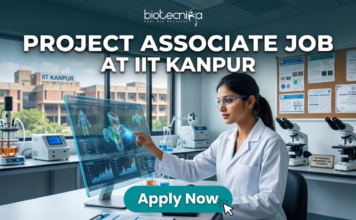 IIT Kanpur Hiring NOW! Project Associate Job Openings in Gene Therapy Project Associate Job at IIT Kanpur Life Science Research 2026. A promotional featured image banner for a 'PROJECT ASSOCIATE JOB AT IIT KANPUR', with the 'biotecnika YOUR BIO RESOURCE' logo at the top left. The image depicts a female scientist in a white lab coat and safety glasses, seated at a desk in a modern laboratory. She is interacting with a large, transparent holographic display, showing detailed 3D molecular structures and data visualizations. In the background, there is a large window with a view of the 'IIT KANPUR' campus building, other scientific equipment like microscopes and centrifuges, and a corkboard with certificates. A prominent, clickable 'Apply Now' button with a cursor arrow is located at the bottom center.