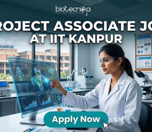Project Associate Job at IIT Kanpur Life Science Research 2026. A promotional featured image banner for a 'PROJECT ASSOCIATE JOB AT IIT KANPUR', with the 'biotecnika YOUR BIO RESOURCE' logo at the top left. The image depicts a female scientist in a white lab coat and safety glasses, seated at a desk in a modern laboratory. She is interacting with a large, transparent holographic display, showing detailed 3D molecular structures and data visualizations. In the background, there is a large window with a view of the 'IIT KANPUR' campus building, other scientific equipment like microscopes and centrifuges, and a corkboard with certificates. A prominent, clickable 'Apply Now' button with a cursor arrow is located at the bottom center.
