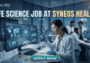 Life Science Job at Syneos Health | Central Monitor Role. A professional promotional graphic for a "Life Science Job at Syneos Health" featuring the Biotecnika logo at the top. The image depicts a female scientist in a white lab coat and blue gloves using a multichannel pipette on a microplate in a modern, high-tech laboratory. A large computer monitor in the foreground displays complex global data visualizations, including a world map with interconnected nodes, cellular diagrams, and data graphs. In the background, other researchers are seen working at their stations. The laboratory features large glass windows that reveal a glowing city skyline at night. A large, prominent "APPLY NOW" button is centered at the bottom.