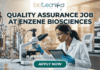 A professional promotional graphic for a Quality Assurance Job at Enzene Biosciences. At the top center is the Biotecnika Professional logo, followed by the headline "QUALITY ASSURANCE JOB AT ENZENE BIOSCIENCES" in bold, dark green lettering. The image features a female scientist in a white lab coat and safety glasses, focused on a digital tablet while working in a high-tech laboratory. The setting includes a microscope, rows of clear vials on a lab bench, and large-scale industrial bioreactors in the background. A glowing teal "APPLY NOW" button is positioned prominently at the bottom center.