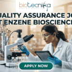 A professional promotional graphic for a Quality Assurance Job at Enzene Biosciences. At the top center is the Biotecnika Professional logo, followed by the headline "QUALITY ASSURANCE JOB AT ENZENE BIOSCIENCES" in bold, dark green lettering. The image features a female scientist in a white lab coat and safety glasses, focused on a digital tablet while working in a high-tech laboratory. The setting includes a microscope, rows of clear vials on a lab bench, and large-scale industrial bioreactors in the background. A glowing teal "APPLY NOW" button is positioned prominently at the bottom center.