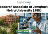 Research Associate Job at JNU Life Science Job 2026. A recruitment banner advertisement for a "Project Associate at Jawaharlal Nehru University (JNU)" position, presented by Biotecnika. At the top center, the Biotecnika "Your Bio Resource" logo is displayed. Below, large black and green text reads, "Project Associate at Jawaharlal Nehru University (JNU)". The main visual at the bottom is a detailed composite photograph blending an extensive, multi-story, red-brick university complex with a modern laboratory setting. In the distance, a sign is visible on a building that reads "IIT KANPUR", creating a text contradiction. On the right, a young woman in a lab coat and safety glasses interacts with a futuristic, transparent table-top display screen showing glowing 3D molecular structures, labeled "DNA STRUCTURE", along with charts and data graphs. In the bottom center, a prominent teal button with a mouse cursor reads "Apply Now", with a small desk plaque next to it that reads "JNU RESEARCH ASSOCIATE PROGRAM". Microscopes and lab equipment are in the background.