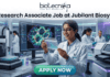 Research Associate Job at Jubilant Biosys | Apply Now. A professional 1280x720 featured image for a job posting titled 'Research Associate Job at Jubilant Biosys.' The image features the Biotecnika logo at the top center and depicts a smiling female scientist in a white lab coat holding a multi-well plate in a high-tech, modern laboratory. The background shows advanced scientific equipment and a large monitor displaying vibrant cellular microscopy. At the bottom, there is a prominent green 'APPLY NOW' button."