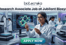Research Associate Job at Jubilant Biosys | Apply Now. A professional 1280x720 featured image for a job posting titled 'Research Associate Job at Jubilant Biosys.' The image features the Biotecnika logo at the top center and depicts a smiling female scientist in a white lab coat holding a multi-well plate in a high-tech, modern laboratory. The background shows advanced scientific equipment and a large monitor displaying vibrant cellular microscopy. At the bottom, there is a prominent green 'APPLY NOW' button."