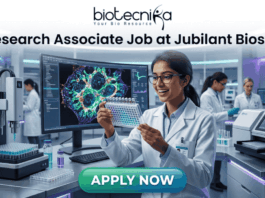 Research Associate Job at Jubilant Biosys | Apply Now. A professional 1280x720 featured image for a job posting titled 'Research Associate Job at Jubilant Biosys.' The image features the Biotecnika logo at the top center and depicts a smiling female scientist in a white lab coat holding a multi-well plate in a high-tech, modern laboratory. The background shows advanced scientific equipment and a large monitor displaying vibrant cellular microscopy. At the bottom, there is a prominent green 'APPLY NOW' button."