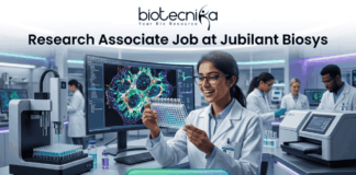 Research Associate Job at Jubilant Biosys –MSc Life Sciences Candidates Apply Today! Research Associate Job at Jubilant Biosys | Apply Now. A professional 1280x720 featured image for a job posting titled 'Research Associate Job at Jubilant Biosys.' The image features the Biotecnika logo at the top center and depicts a smiling female scientist in a white lab coat holding a multi-well plate in a high-tech, modern laboratory. The background shows advanced scientific equipment and a large monitor displaying vibrant cellular microscopy. At the bottom, there is a prominent green 'APPLY NOW' button."