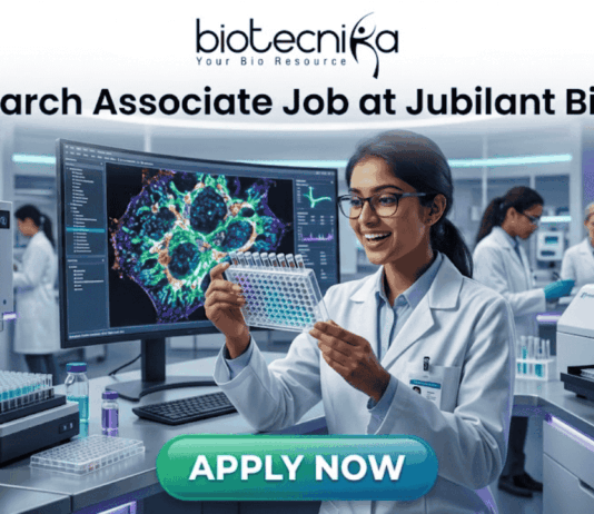 Research Associate Job at Jubilant Biosys | Apply Now. A professional 1280x720 featured image for a job posting titled 'Research Associate Job at Jubilant Biosys.' The image features the Biotecnika logo at the top center and depicts a smiling female scientist in a white lab coat holding a multi-well plate in a high-tech, modern laboratory. The background shows advanced scientific equipment and a large monitor displaying vibrant cellular microscopy. At the bottom, there is a prominent green 'APPLY NOW' button."