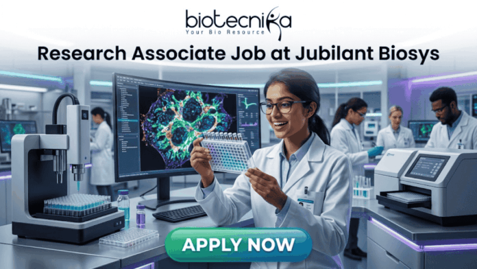 Research Associate Job at Jubilant Biosys –MSc Life Sciences Candidates Apply Today! Research Associate Job at Jubilant Biosys | Apply Now. A professional 1280x720 featured image for a job posting titled 'Research Associate Job at Jubilant Biosys.' The image features the Biotecnika logo at the top center and depicts a smiling female scientist in a white lab coat holding a multi-well plate in a high-tech, modern laboratory. The background shows advanced scientific equipment and a large monitor displaying vibrant cellular microscopy. At the bottom, there is a prominent green 'APPLY NOW' button."