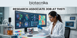 Research Associate Job at THSTI PhD Life Sciences Candidates Earn Upto ₹47,000. A realistic, high-tech featured image for a "Research Associate Job at THSTI." At the top is the Biotecnika logo. The center features a female scientist in a white lab coat and safety glasses, meticulously using a pipette over a multi-well plate. Behind her, a large curved monitor displays complex molecular structures and colorful genomic data charts. The modern laboratory is filled with advanced scientific equipment like a bioreactor and centrifuge. At the bottom, there is a prominent teal "APPLY NOW" button.