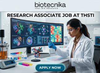 Research Associate Job at THSTI | PhD Life Sciences Candidates Earn Upto ₹47,000 Research Associate Job at THSTI PhD Life Sciences Candidates Earn Upto ₹47,000. A realistic, high-tech featured image for a "Research Associate Job at THSTI." At the top is the Biotecnika logo. The center features a female scientist in a white lab coat and safety glasses, meticulously using a pipette over a multi-well plate. Behind her, a large curved monitor displays complex molecular structures and colorful genomic data charts. The modern laboratory is filled with advanced scientific equipment like a bioreactor and centrifuge. At the bottom, there is a prominent teal "APPLY NOW" button.
