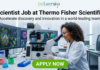 Scientist Job at Thermo Fisher Scientific | High-Paying R&D Role. A professional featured image for a "Scientist Job at Thermo Fisher Scientific." The visual shows a female scientist in a white lab coat, smiling and working with a microscope in a high-tech laboratory. In the background, large computer monitors display colorful, high-resolution cellular imaging. The Biotecnika logo is positioned at the top, and a prominent green "APPLY NOW" button is at the bottom center.