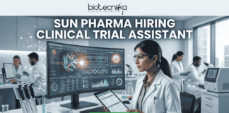 Sun Pharma Hiring Clinical Trial Assistant | Apply for Clinical Research Job! Sun Pharma Hiring Clinical Trial Assistant Apply Now. A professional and high-tech promotional image for a job opening. At the top, the Biotecnika logo is placed above the bold, capitalized headline: "SUN PHARMA HIRING CLINICAL TRIAL ASSISTANT". The central focus is a female scientist in a white lab coat and safety glasses, intently working at a desk. She is looking at a large, curved digital monitor that displays intricate clinical trial data, 3D molecular structures, and a glowing sun-like graphic representing the company theme. The laboratory setting is bright and modern, with other researchers working in the background and scientific equipment like pipettes and vials visible on the desk. At the bottom center, a vibrant green "APPLY NOW" button stands out, making the image an effective, scroll-stopping recruitment graphic.