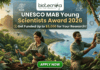 UNESCO MAB Young Scientists Award 2026 – Get Funded Up to $5,000 for Your Research! A featured job post image for the UNESCO MAB Young Scientists Award 2026. At the top center is the Biotecnika logo. The main text reads "UNESCO MAB Young Scientists Award 2026" with a sub-headline, "Get Funded Up to $5,000 for Your Research!" The background features a realistic and vibrant tropical coastal landscape with lush green mountains and turquoise water. Two scientists—a woman and a man in khaki field gear—are shown conducting environmental research; the woman uses a handheld probe and tablet while the man inspects a sensor device mounted on a tree. Drones fly in the background, emphasizing a high-tech conservation theme. At the bottom center is a prominent green "APPLY NOW" button.