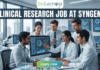 Urgent Hiring! Clinical Research Job at Syngene – Apply Today! A featured image for a job page, part of Biotecnika, showcasing a clinical research position at Syngene. At the top center is the Biotecnika logo, with the text 'Your Bio Resource'. The main bold title is 'CLINICAL RESEARCH JOB AT SYNGENE'. The background features a detailed, realistic photograph of a diverse team of five scientists and professionals (two women, three men) collaborating in a modern, well-equipped laboratory, looking collaboratively into a computer monitor showing data charts, graphs, and a detailed 3D molecular structure. The lab has advanced scientific instruments, beakers, and shelves, visible next to a large window. The overall scene conveys scientific innovation, teamwork, and professionalism. At the bottom center, there is a prominent orange rectangular button labeled 'Apply now'.