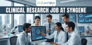 Urgent Hiring! Clinical Research Job at Syngene – Apply Today! A featured image for a job page, part of Biotecnika, showcasing a clinical research position at Syngene. At the top center is the Biotecnika logo, with the text 'Your Bio Resource'. The main bold title is 'CLINICAL RESEARCH JOB AT SYNGENE'. The background features a detailed, realistic photograph of a diverse team of five scientists and professionals (two women, three men) collaborating in a modern, well-equipped laboratory, looking collaboratively into a computer monitor showing data charts, graphs, and a detailed 3D molecular structure. The lab has advanced scientific instruments, beakers, and shelves, visible next to a large window. The overall scene conveys scientific innovation, teamwork, and professionalism. At the bottom center, there is a prominent orange rectangular button labeled 'Apply now'.