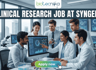 Urgent Hiring! Clinical Research Job at Syngene – Apply Today! Urgent Hiring! Clinical Research Job at Syngene – Apply Today! A featured image for a job page, part of Biotecnika, showcasing a clinical research position at Syngene. At the top center is the Biotecnika logo, with the text 'Your Bio Resource'. The main bold title is 'CLINICAL RESEARCH JOB AT SYNGENE'. The background features a detailed, realistic photograph of a diverse team of five scientists and professionals (two women, three men) collaborating in a modern, well-equipped laboratory, looking collaboratively into a computer monitor showing data charts, graphs, and a detailed 3D molecular structure. The lab has advanced scientific instruments, beakers, and shelves, visible next to a large window. The overall scene conveys scientific innovation, teamwork, and professionalism. At the bottom center, there is a prominent orange rectangular button labeled 'Apply now'.