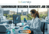 IIT Gandhinagar Research Associate Job 2026 | Get Upto ₹54,000 Salary | Apply Before Deadline! A professional promotional image for a Research Associate job at IIT Gandhinagar in 2026. At the top, the Biotecnika Global logo is centered. Large, bold text reads "IIT GANDHINAGAR RESEARCH ASSOCIATE JOB 2026". In the center, a female scientist wearing a white lab coat and safety glasses is focused on her work, using a micropipette to dispense liquid into a multi-well plate. She sits in a modern, well-equipped laboratory with advanced research equipment and computer monitors displaying molecular data. Large windows in the background reveal a bright, green university campus. At the bottom, a prominent "Apply Now" button with an orange-to-green gradient is displayed.