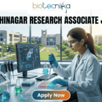 A professional promotional image for a Research Associate job at IIT Gandhinagar in 2026. At the top, the Biotecnika Global logo is centered. Large, bold text reads "IIT GANDHINAGAR RESEARCH ASSOCIATE JOB 2026". In the center, a female scientist wearing a white lab coat and safety glasses is focused on her work, using a micropipette to dispense liquid into a multi-well plate. She sits in a modern, well-equipped laboratory with advanced research equipment and computer monitors displaying molecular data. Large windows in the background reveal a bright, green university campus. At the bottom, a prominent "Apply Now" button with an orange-to-green gradient is displayed.