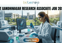 IIT Gandhinagar Research Associate Job 2026 | Get Upto ₹54,000 Salary | Apply Before Deadline! A professional promotional image for a Research Associate job at IIT Gandhinagar in 2026. At the top, the Biotecnika Global logo is centered. Large, bold text reads "IIT GANDHINAGAR RESEARCH ASSOCIATE JOB 2026". In the center, a female scientist wearing a white lab coat and safety glasses is focused on her work, using a micropipette to dispense liquid into a multi-well plate. She sits in a modern, well-equipped laboratory with advanced research equipment and computer monitors displaying molecular data. Large windows in the background reveal a bright, green university campus. At the bottom, a prominent "Apply Now" button with an orange-to-green gradient is displayed.