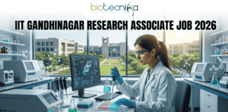 IIT Gandhinagar Research Associate Job 2026 | Get Upto ₹54,000 Salary | Apply Before Deadline! A professional promotional image for a Research Associate job at IIT Gandhinagar in 2026. At the top, the Biotecnika Global logo is centered. Large, bold text reads "IIT GANDHINAGAR RESEARCH ASSOCIATE JOB 2026". In the center, a female scientist wearing a white lab coat and safety glasses is focused on her work, using a micropipette to dispense liquid into a multi-well plate. She sits in a modern, well-equipped laboratory with advanced research equipment and computer monitors displaying molecular data. Large windows in the background reveal a bright, green university campus. At the bottom, a prominent "Apply Now" button with an orange-to-green gradient is displayed.
