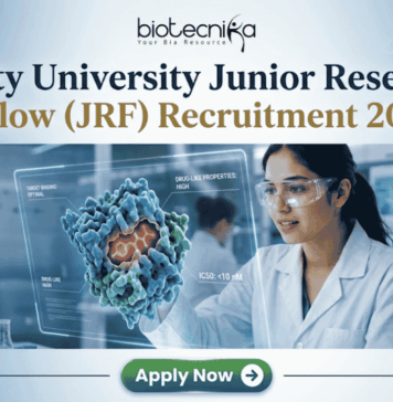 Amity University Junior Research Fellow (JRF) Recruitment 2026 | Apply Now Amity University Junior Research Fellow (JRF) Recruitment 2026 Apply Now. A featured image for a job posting titled "Amity University Junior Research Fellow (JRF) Recruitment 2026." At the top center is the Biotecnika logo. Below it, the main heading reads "Amity University Junior Research Fellow (JRF) Recruitment 2026" in a bold, professional font. The background features a high-tech, modern laboratory where a female researcher in a white lab coat and safety goggles is intensely focused on a glowing, 3D holographic molecular model representing drug research. The lab is clean and corporate, with various scientific equipment visible in soft focus. At the bottom center, there is a prominent green "Apply Now" button with a white arrow icon. The overall color palette is a professional mix of blue, white, and teal, designed to be eye-catching and authoritative.