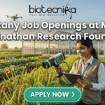 Botany Job Openings at M.S. Swaminathan Research Foundation (MSSRF) in Kerala Botany Job Openings at M.S. Swaminathan Research Foundation (MSSRF) in Kerala Apply Now. A featured job banner image with the bold text "Botany Job Openings at M.S. Swaminathan Research Foundation" at the center and a prominent green "APPLY NOW >" button below it. The image is a photo-realistic scene of a woman researcher in a sunlit agricultural field, holding a hand-held analytical device and a tablet, looking at the tablet with a smile. Behind her are lush crop rows (like rice paddy), a large modern greenhouse, and a drone in the air, all bathed in warm golden hour light. The Biotecnika logo is at the very top.