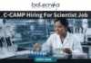 C-CAMP Hiring For Scientist Job | Fresh PhD Candidates Apply Now C-CAMP Hiring For Scientist Job Fresh PhD Candidates Apply Now. A job advertisement banner from Biotecnika, with the main title at the top, "C-CAMP Hiring For Scientist Job." The Biotecnika logo with the text "Your Bio Resource" is in the top center. The visual focuses on a female scientist in a modern drug discovery laboratory, wearing safety glasses and a lab coat. She is looking at a large, holographic-style transparent digital display that shows complex scientific information. This includes detailed 2D molecular structures under the heading "Compound Library Screen," a high-throughput screening assay graph, and text identifying the "Target Protein: SARS-CoV-2 M^pro." The lab environment around her is fully equipped with advanced automated systems, analytical machines, multi-well plates, vials, and a prominent rack of sophisticated pipettes. At the bottom center, there is a large, prominent blue button with the text "APPLY NOW >."