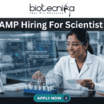 C-CAMP Hiring For Scientist Job | Fresh PhD Candidates Apply Now C-CAMP Hiring For Scientist Job Fresh PhD Candidates Apply Now. A job advertisement banner from Biotecnika, with the main title at the top, "C-CAMP Hiring For Scientist Job." The Biotecnika logo with the text "Your Bio Resource" is in the top center. The visual focuses on a female scientist in a modern drug discovery laboratory, wearing safety glasses and a lab coat. She is looking at a large, holographic-style transparent digital display that shows complex scientific information. This includes detailed 2D molecular structures under the heading "Compound Library Screen," a high-throughput screening assay graph, and text identifying the "Target Protein: SARS-CoV-2 M^pro." The lab environment around her is fully equipped with advanced automated systems, analytical machines, multi-well plates, vials, and a prominent rack of sophisticated pipettes. At the bottom center, there is a large, prominent blue button with the text "APPLY NOW >."