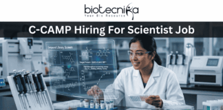 C-CAMP Hiring For Scientist Job | Fresh PhD Candidates Apply Now C-CAMP Hiring For Scientist Job Fresh PhD Candidates Apply Now. A job advertisement banner from Biotecnika, with the main title at the top, "C-CAMP Hiring For Scientist Job." The Biotecnika logo with the text "Your Bio Resource" is in the top center. The visual focuses on a female scientist in a modern drug discovery laboratory, wearing safety glasses and a lab coat. She is looking at a large, holographic-style transparent digital display that shows complex scientific information. This includes detailed 2D molecular structures under the heading "Compound Library Screen," a high-throughput screening assay graph, and text identifying the "Target Protein: SARS-CoV-2 M^pro." The lab environment around her is fully equipped with advanced automated systems, analytical machines, multi-well plates, vials, and a prominent rack of sophisticated pipettes. At the bottom center, there is a large, prominent blue button with the text "APPLY NOW >."