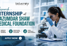 Research Internship at Mazumdar Shaw Medical Foundation | Life Science Post Graduates Must Apply! Research Internship at Mazumdar Shaw Medical Foundation. Research Internship at Mazumdar Shaw Medical Foundation banner featuring a scientist using a microscope in a modern lab, Biotecnika logo at the top, and an ‘Apply Now’ button highlighting hands-on research opportunities.