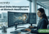 Clinical Data Management Job at Biotech Healthcare | Apply Online Here Clinical Data Management Job at Biotech Healthcare Apply Online Here. A corporate job advertisement for a "Clinical Data Management Job at Biotech Healthcare," featuring a professional female of South Asian descent smiling and working at a desk with dual monitors. The screens display data visualizations of molecular structures and CDM progress dashboards with text labels like "CDM QUERY RESOLUTION" and "VALIDATED DATA STREAM." Through the large office window behind her, a modern building labeled "HOSPITAL" and "CLINICAL RESEARCH CENTRE" is visible. The Biotecnika logo is at the top left, and a prominent green "APPLY NOW" button with an arrow icon is at the bottom right. Other colleagues are working blurred in the background.
