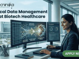 Clinical Data Management Job at Biotech Healthcare | Apply Online Here Clinical Data Management Job at Biotech Healthcare Apply Online Here. A corporate job advertisement for a "Clinical Data Management Job at Biotech Healthcare," featuring a professional female of South Asian descent smiling and working at a desk with dual monitors. The screens display data visualizations of molecular structures and CDM progress dashboards with text labels like "CDM QUERY RESOLUTION" and "VALIDATED DATA STREAM." Through the large office window behind her, a modern building labeled "HOSPITAL" and "CLINICAL RESEARCH CENTRE" is visible. The Biotecnika logo is at the top left, and a prominent green "APPLY NOW" button with an arrow icon is at the bottom right. Other colleagues are working blurred in the background.