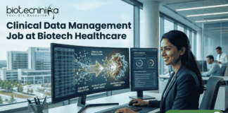 Clinical Data Management Job at Biotech Healthcare Apply Online Here. A corporate job advertisement for a "Clinical Data Management Job at Biotech Healthcare," featuring a professional female of South Asian descent smiling and working at a desk with dual monitors. The screens display data visualizations of molecular structures and CDM progress dashboards with text labels like "CDM QUERY RESOLUTION" and "VALIDATED DATA STREAM." Through the large office window behind her, a modern building labeled "HOSPITAL" and "CLINICAL RESEARCH CENTRE" is visible. The Biotecnika logo is at the top left, and a prominent green "APPLY NOW" button with an arrow icon is at the bottom right. Other colleagues are working blurred in the background.