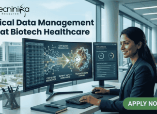 Clinical Data Management Job at Biotech Healthcare | Apply Online Here Clinical Data Management Job at Biotech Healthcare Apply Online Here. A corporate job advertisement for a "Clinical Data Management Job at Biotech Healthcare," featuring a professional female of South Asian descent smiling and working at a desk with dual monitors. The screens display data visualizations of molecular structures and CDM progress dashboards with text labels like "CDM QUERY RESOLUTION" and "VALIDATED DATA STREAM." Through the large office window behind her, a modern building labeled "HOSPITAL" and "CLINICAL RESEARCH CENTRE" is visible. The Biotecnika logo is at the top left, and a prominent green "APPLY NOW" button with an arrow icon is at the bottom right. Other colleagues are working blurred in the background.