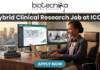 Hybrid Clinical Research Job at ICON | Life Science Graduates Must Apply! Hybrid Clinical Research Job at ICON | Life Science Graduates Must Apply! A job advertisement banner from Biotecnika for a Clinical Research position at ICON. The top center features the "biotecnika" logo with the tagline "Your Bio Resource". Below the logo, a prominent dark grey bar contains the large, bold white text headline: "Hybrid Clinical Research Job at ICON". The main visual is a detailed, realistic illustrative scene of a professional woman in a blazer and headphones seated at a desk with an office cityscape in the background. She is looking at three computer monitors: one showing a "Global Trial Management" data dashboard, another showing a live video conference with a team (labeled "Team Check-in - Remote Site"), and the third is a laptop. Objects on the desk include a coffee mug that reads "COFFEE & CLINICAL DATA", a stack of "Good Clinical Practice Guide" books, and vials. At the bottom center, a pulsing teal-colored rounded rectangular button with white text reads "APPLY NOW".